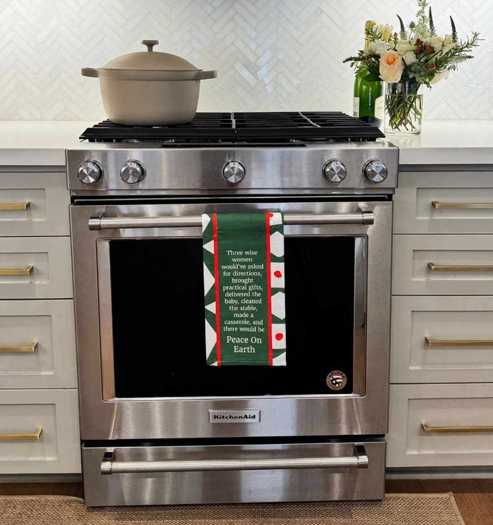 A stainless steel KitchenAid oven with a Drinks On Me CHRISTMAS Peace on Earth Dish Towel hanging on the handle, a beige pot on the stovetop, and a vase of flowers on a white countertop in a modern kitchen.
