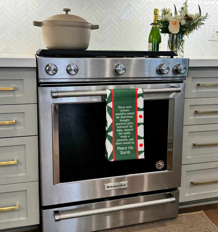 A modern stainless steel oven with a Drinks On Me CHRISTMAS Peace on Earth Dish Towel hanging on the handle, a beige pot on the stovetop, a bottle of sparkling wine, and flowers on the countertop.