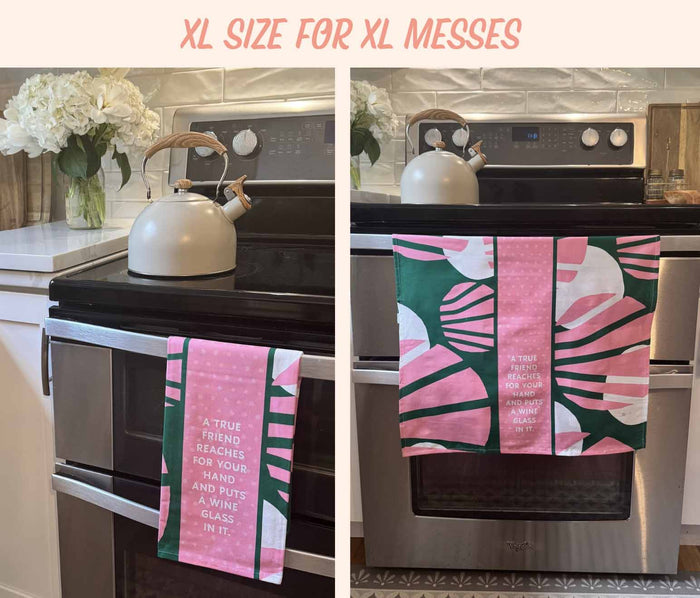 Two photos of a kitchen stove showcase the "Young and Fun Dish Towel" by Drinks On Me. The towel features pink and green abstract patterns with a witty wine-and-friendship quote. A white kettle sits on the stove, with fresh white flowers on the counter.