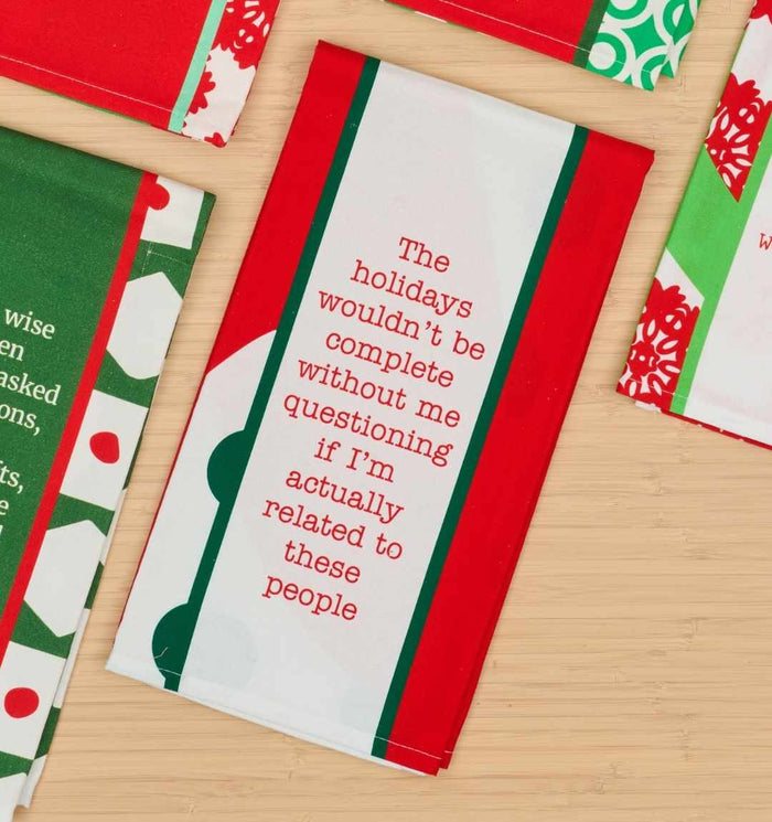 The Drinks On Me CHRISTMAS Related Dish Towel features festive red, green, and white colors with a sarcastic holiday saying—a perfect humorous kitchen gift. Shown on a light wood surface.