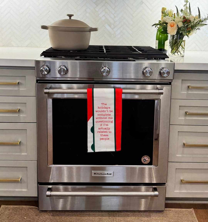 A stainless steel oven with a beige pot on top and a Drinks On Me CHRISTMAS Related Dish Towel hanging from the handle, set in a modern kitchen with white cabinets, herringbone backsplash, and a vase of flowers.