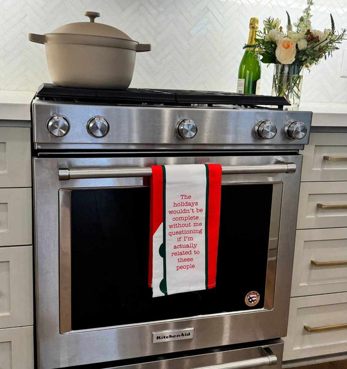 A modern stainless steel oven displays the Drinks On Me CHRISTMAS Related Dish Towel—a red and white humorous gift reading, “The holidays wouldn’t be complete without me questioning if I’m actually related to these people.” A pot and champagne bottle sit above.