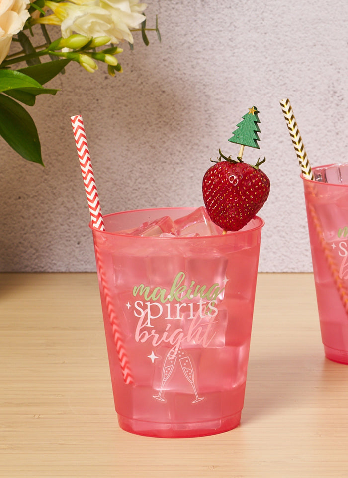 A pink cup with “making spirits bright” and champagne glasses, filled with ice, a red-striped straw, and a strawberry topped with Simply Serv'd Christmas Tree Large Cocktail Picks (30pk). Flowers and another cup add festive cheer in the background.