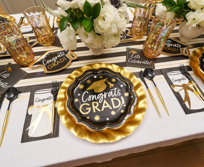 A festive graduation table is set with Sophistiplate Classic Grad Dinner Plates (8pk), gold utensils, a striped tablecloth, floral centerpieces, plus celebratory cards and glasses.
