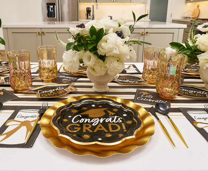 A graduation table is set with Sophistiplate's Classic Grad Dinner Plates (8pk) featuring "Congrats Grad," gold cutlery, amber glasses, white floral centerpieces, themed napkins, and a striped runner in a modern kitchen.