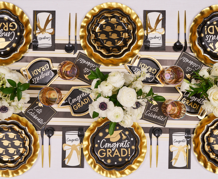 Celebrate with the Classic Grad Table Setting by Sophistiplate—designer black, gold, and white plates, Congrats Grad! and Let's Celebrate! napkins, gold cutlery, floral centerpieces, and a striped runner for a festive graduation party.