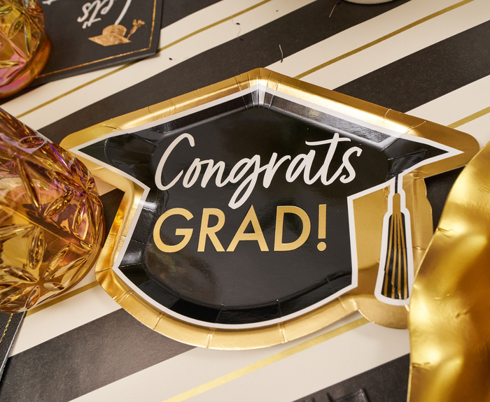 The Sophistiplate Classic Grad Table Setting features a black and gold graduation cap-shaped plate with "Congrats GRAD!" on a black and white striped table, surrounded by festive graduation party supplies and decor.