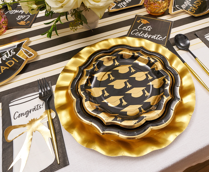A festive graduation table with Sophistiplate Classic Grad Salad Plates (8pk), gold and black party plates, "Congrats!" and "Let's Celebrate!" napkins, gold utensils, disposable salad plates, and a white flower centerpiece.