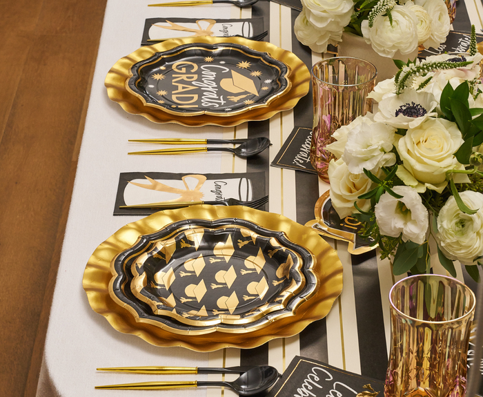 The Classic Grad Table Setting by Sophistiplate features designer plates with graduation cap motifs, gold utensils, clear ribbed glasses, floral centerpieces, and congratulatory cards—perfect for a stylish graduation celebration.
