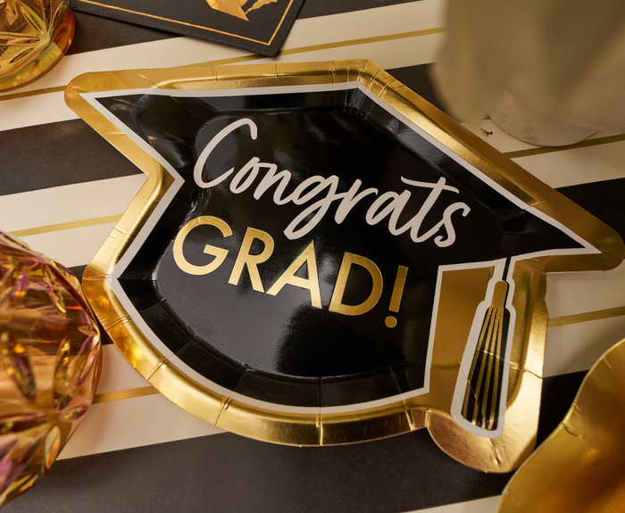 The Sophistiplate Classic Grad Table Setting features a designer plate shaped like a graduation cap with “Congrats GRAD!” in white and gold, set on black and white stripes—an ideal choice for your graduation party supplies.