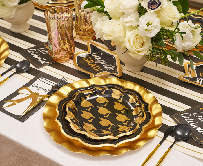 Celebrate in style with the Classic Grad Table Setting by Sophistiplate—designer plates with graduation caps, gold utensils, a striped runner, white florals, gold glasses, and "Congrats Grad" decor. The perfect disposable supplies for your graduation party!.