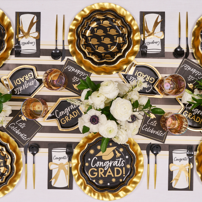 The Sophistiplate Classic Grad Table Setting features black and gold paper plates, “Congrats Grad!” signs, gold utensils, bow-tied napkins, and white floral centerpieces—perfect for stylish graduation party decor.