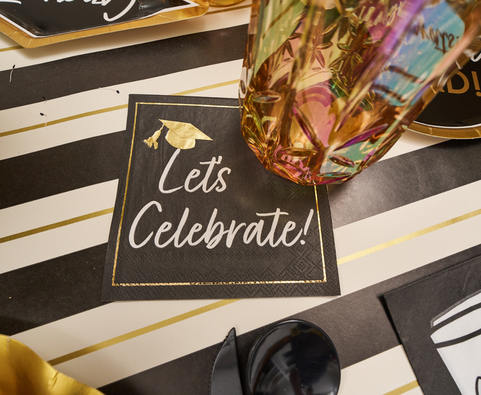 A close-up of the Classic Grad Table Setting by Sophistiplate: a black napkin with gold graduation cap and "Let's Celebrate!" on a striped table, paired with patterned glass, black utensils, and stylish graduation party supplies.