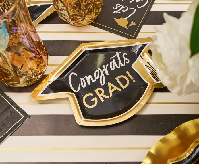 The Sophistiplate Classic Grad Table Setting features a grad cap-shaped plate with “Congrats GRAD!”, gold-rimmed glasses, a black napkin, and white flowers—ideal for an elegant, disposable graduation celebration tablescape.