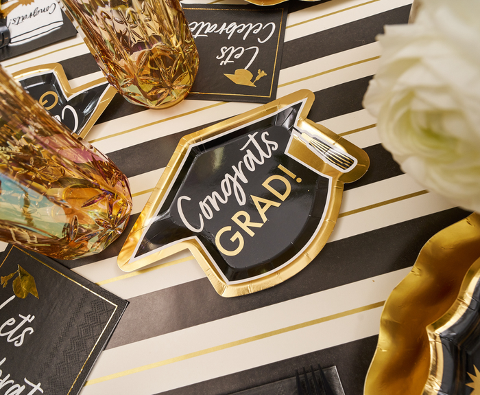 The Sophistiplate Classic Grad Table Setting features a black and gold graduation cap-shaped plate with "Congrats GRAD!", gold-rimmed glasses, napkins, and white flowers on a striped tablecloth.