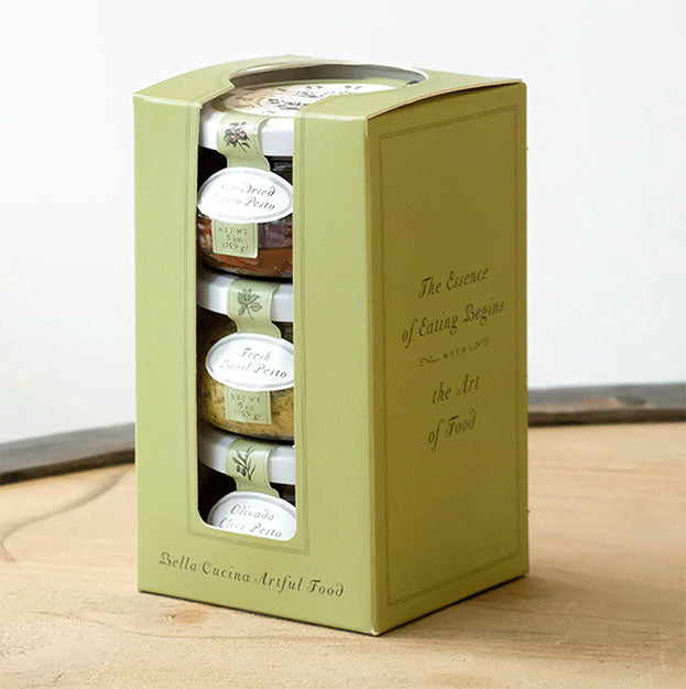 A green box labeled "Classic Pesto Collection Trio" by Bella Cucina holds three stacked jars of pesto and features the phrase "The Essence of Eating Begins with the Art of Food" on the side, set atop a wooden surface.