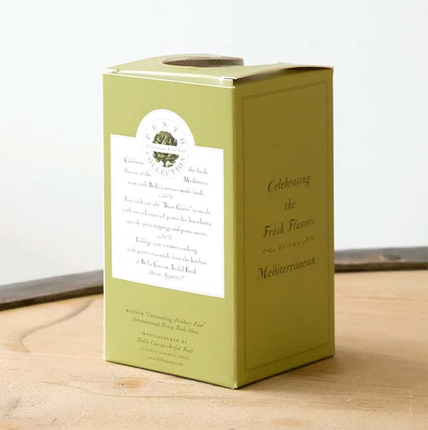 A green rectangular box from Bella Cucina, labeled "Classic Pesto Collection Trio" and featuring a circular emblem, sits on a wooden surface. Printed text highlights the fresh flavors of the Mediterranean, hinting at a delicious pesto trio inside.