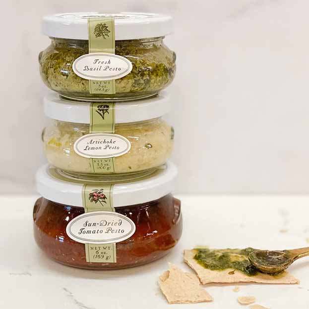 The Bella Cucina Classic Pesto Collection Trio—Fresh Basil, Artichoke Lemon, and Sun-Dried Tomato—are stacked on a white surface beside crackers topped with basil pesto and a small spoon.