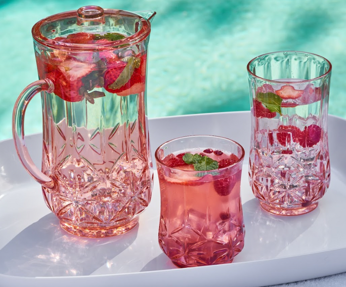 A Sophistiplate Classic Pitcher in Blush (53 Oz) and two glasses with fruity drinks, garnished with strawberries, raspberries, and mint, rest on a white tray by a bright blue pool.