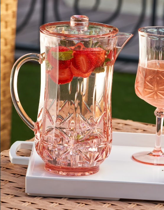 A Sophistiplate Classic Pitcher in Blush (53 oz), BPA-free, filled with water, strawberries, and mint sits on a white tray next to a matching pink drink outdoors on a wicker table.
