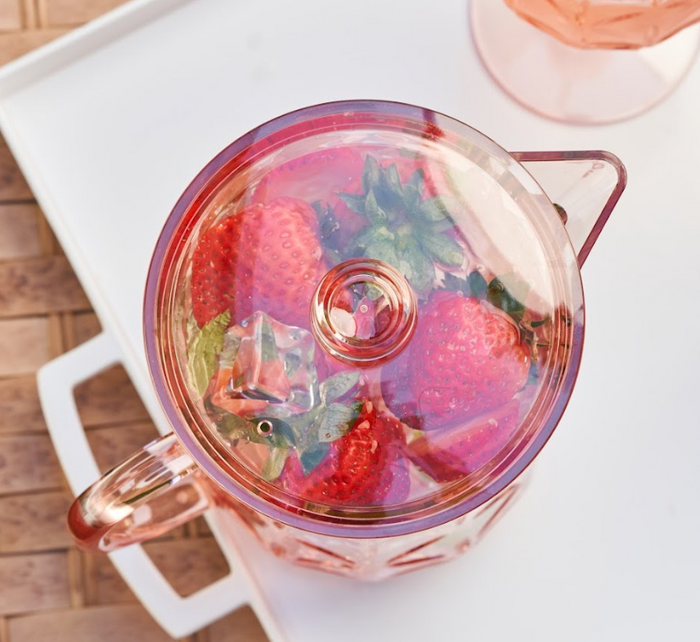 The Sophistiplate Classic Pitcher | Blush | 53 Oz, filled with fresh strawberries, sits on a white tray atop a woven surface. A matching pink glass peeks from the corner, all viewed from above.