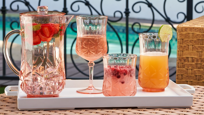 The Sophistiplate Classic Pitcher in Blush (53 oz), dishwasher safe and shatterproof, stands by a pool with decorative railing, filled with colorful drinks in three glasses, each garnished with fruit slices and berries.