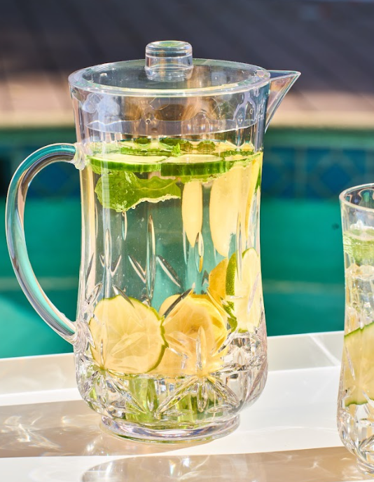 A Sophistiplate Classic Pitcher | Clear | 53 Oz, filled with water, lime slices, and mint leaves, sits on a white surface near a swimming pool in bright sunlight.
