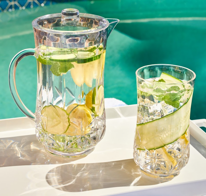 A Sophistiplate Classic Pitcher (Clear, 53 Oz) and a glass filled with ice water, lemon, cucumber, and mint sit on a white tray by the pool in bright sunlight.