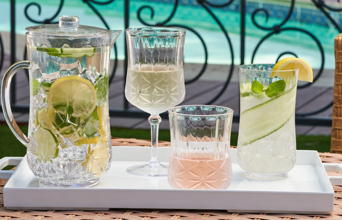 A tray by the pool holds a glass pitcher of citrus-mint water, a Sophistiplate Wine Classic Stemmed | Clear | 16 Oz with a light drink, a tumbler with pink beverage, and a tall glass of lemon-cucumber water.