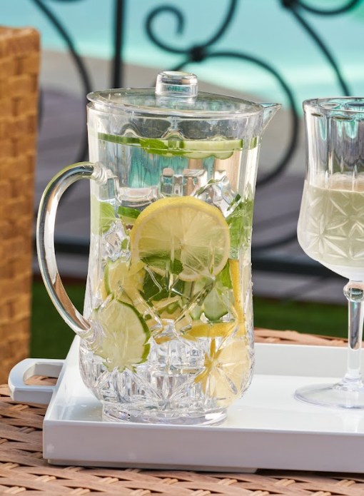 A Sophistiplate Classic Pitcher | Clear | 53 Oz, filled with ice, lemon slices, and mint leaves, sits on a white tray beside a glass, with a blurred pool and decorative railing in the background.