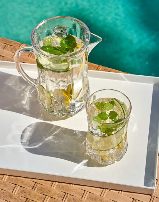 The Sophistiplate Classic Pitcher (Clear, 53 Oz) and a glass with water, lemon slices, and mint sit on a white tray next to a woven surface, with bright sunlight and a blue pool in the background.