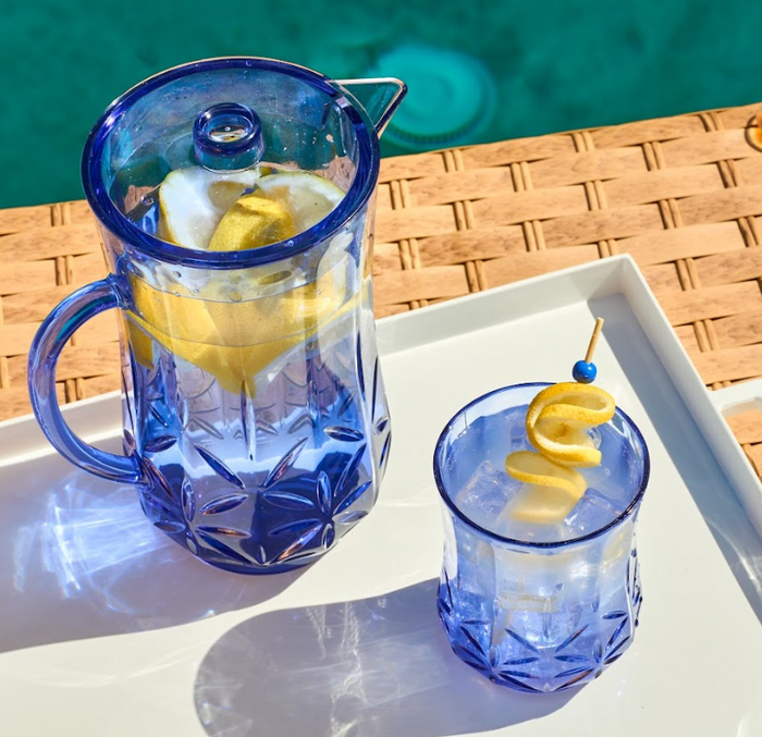The Sophistiplate Classic Pitcher in Cobalt (53 Oz) and a matching glass, both with lemon slices and ice, sit on a white tray by the pool, their refreshing look enhanced by sunlight.