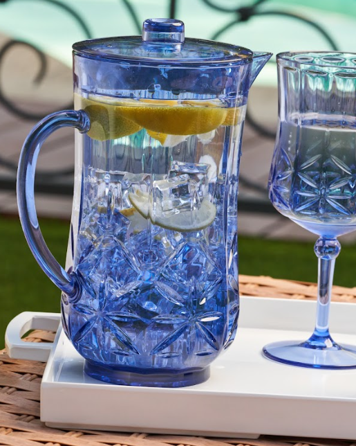 A Sophistiplate Classic Pitcher in Cobalt (53 Oz), BPA-free, filled with ice, lemon slices, and water sits on a white tray beside a matching blue glass against a blurred outdoor background.