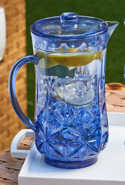 The Sophistiplate Classic Pitcher | Cobalt | 53 Oz, filled with ice water and lemon slices, sits on a white tray outdoors against a green lawn and brick wall.