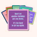A stack of colorful Drinks On Me Coaster Bundle: Birthday cards with funny messages, featuring a purple card that says: Don't let another birthday get you down. It’s too hard to get up again—perfect for friends or your coaster collection.