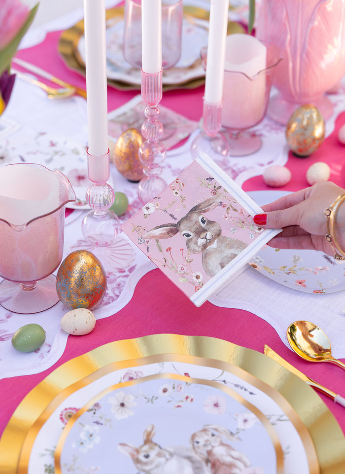 A hand holds a pink bunny napkin over the Ultimate Easter Tablescape for 16: Charming Easter by Sophistiplate LLC, set with gold-trimmed plates, pastel candles, decorative eggs, and pink glassware on a pink and white tablecloth.