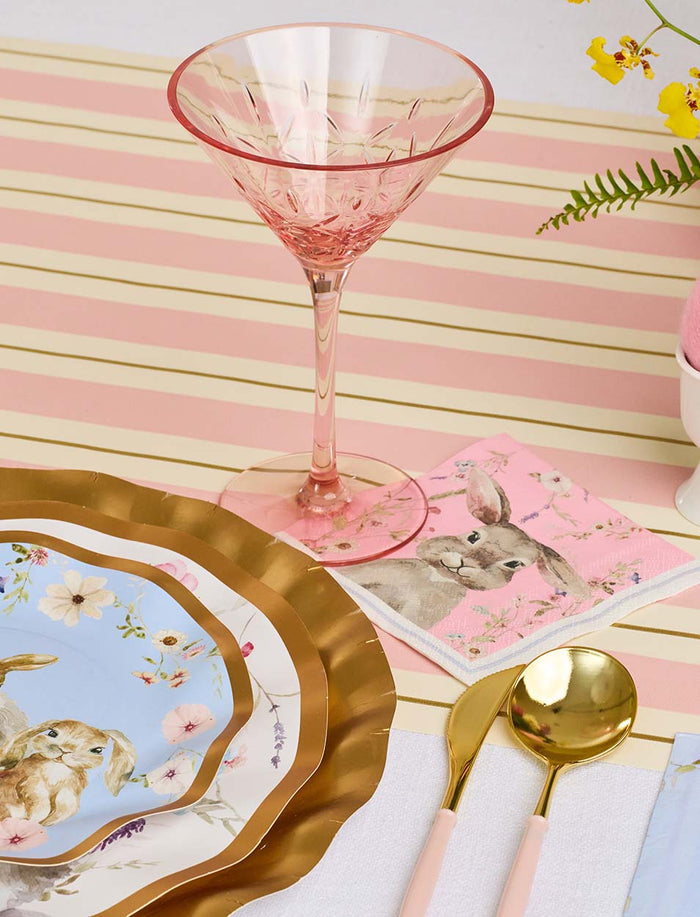 A pink martini glass sits on a striped placemat with gold and pink utensils, brunch tableware, and a Sophistiplate Cocktail Napkin Charming Easter—featuring a rabbit illustration and flowers—from the 26-piece package.
