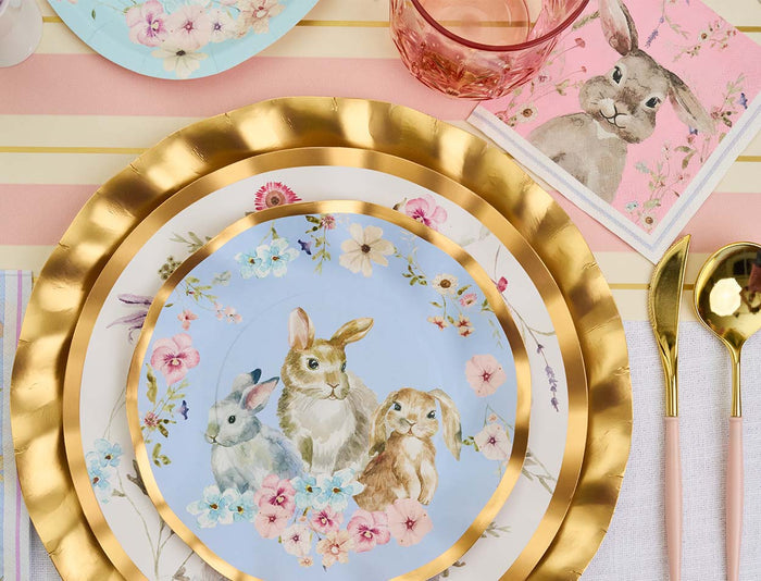 A brunch table is set with gold-rimmed plates, floral bunny plates, gold utensils, a pink glass, and Sophistiplate's Cocktail Napkin Charming Easter (26/pkg) featuring a rabbit on pink. Pink and white stripes cover the tablecloth.