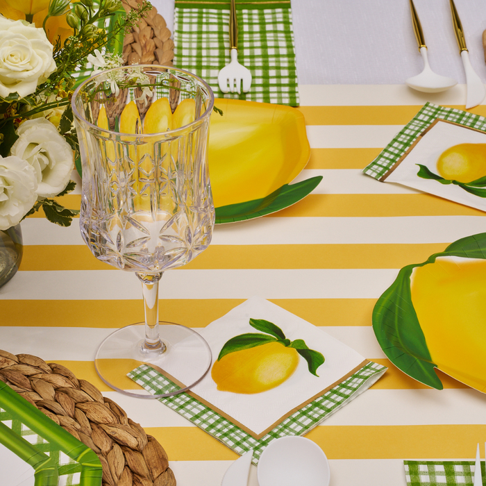 A table set for a meal features a yellow and white striped tablecloth, Lemon and Stem Collection plates, Sophistiplate's Cocktail Napkin Lemon and Stem 3 Ply/20pk, a crystal glass, gold utensils, and a white rose and greenery centerpiece.