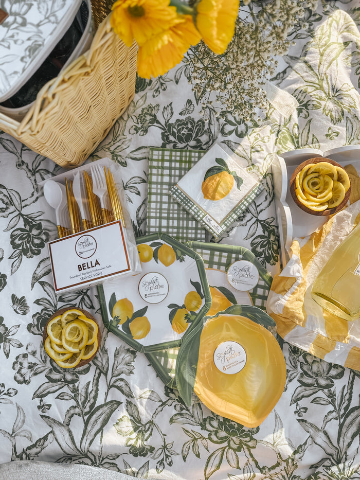 A picnic on a floral blanket with yellow flowers, Sophistiplate Guest Towel Lemon and Stem 3 Ply/20pk napkins, yellow utensils, plates, a wicker basket, and a container of lemon slices creates a bright, cheerful scene.