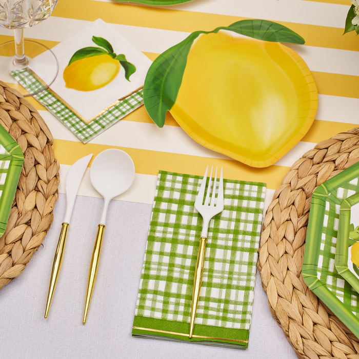 A table set with woven placemats, green plaid napkins, gold and white cutlery, lemon-shaped plates, and Sophistiplate's Lemon and Stem 3 Ply Cocktail Napkins (20pk) on a yellow and white striped tablecloth.