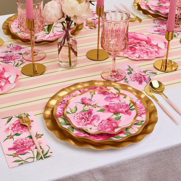 A table set with gold-edged plates and cutlery, pink floral plates, Sophistiplate Cocktail Napkin Pink Peonies (26/pkg), pink candles in gold holders, crystal glasses, all atop a white cloth with a pink and gold striped runner.