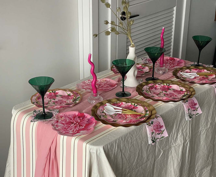 A dining table set with Sophistiplate's Cocktail Napkin Pink Peonies, floral plates, green glasses, striped and pink runners, wavy candles, and a white vase with dried flowers creates an elegant, festive mood.