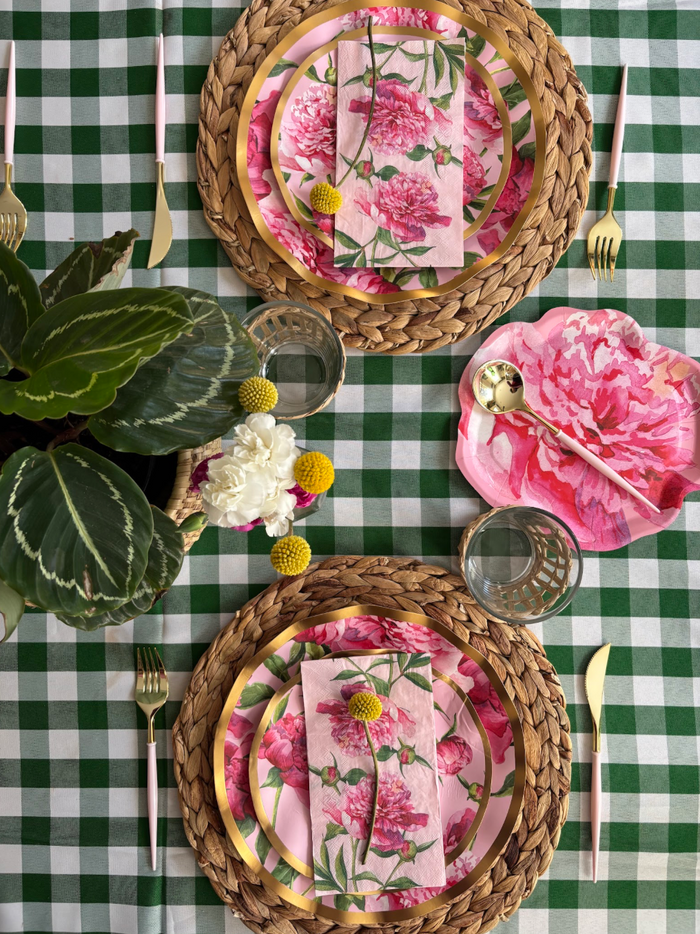 A table set with Sophistiplate’s Cocktail Napkin Pink Peonies (26/pkg), woven placemats, floral pink plates, gold cutlery, and glassware atop a green-and-white checkered tablecloth features a potted plant and pink peonies as the centerpiece.