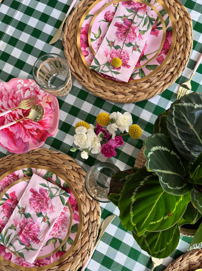 A spring table set with Sophistiplate's Cocktail Napkin Pink Peonies (26pk), floral plates, gold cutlery, woven placemats, and glasses on a green checkered tablecloth, accented by pink peonies and greenery.