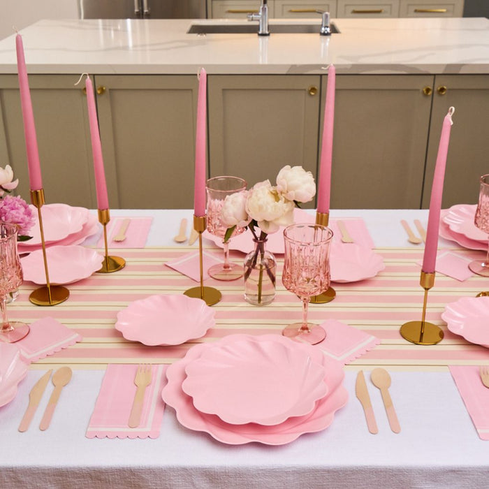 The Eco-Friendly Table Setting- Blush by Sophistiplate LLC features blush scalloped plates, pink napkins, wooden cutlery, pink glassware, gold candle holders with tall pink candles, and small white floral vases on a white tablecloth.