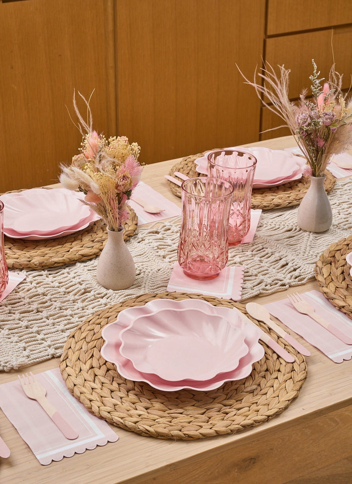 The Eco-Friendly Table Setting-Blush by Sophistiplate LLC features disposable plates, pink glasses, woven placemats, pastel napkins, wooden cutlery, and dried flower vases on a crocheted runner—perfect for a table set for four. Light wood furniture completes the scene.