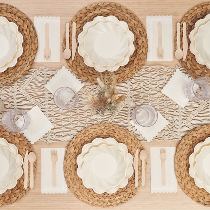 The Eco-Friendly Table Setting- Cream by Sophistiplate features woven placemats, scalloped white compostable plates, wooden cutlery, clear glasses, white napkins, a macramé runner, and a rustic floral centerpiece.