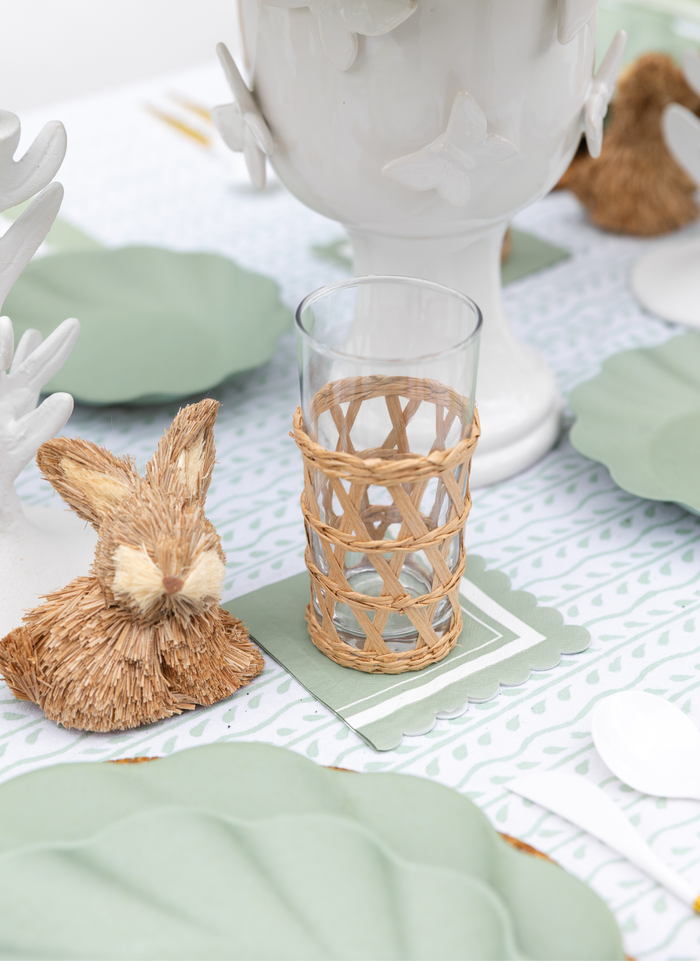 The Eco-friendly Table Setting- Sage by Sophistiplate LLC features a woven straw bunny ornament, glass in a wicker holder on a green coaster, white utensils, compostable leaf-shaped plates, and a white butterfly pitcher.