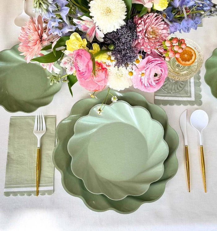 The Eco-friendly Table Setting- Sage by Sophistiplate LLC features pastel green scalloped compostable plates with gold and white cutlery, arranged with a green napkin and drink, and a vibrant bouquet at the center.